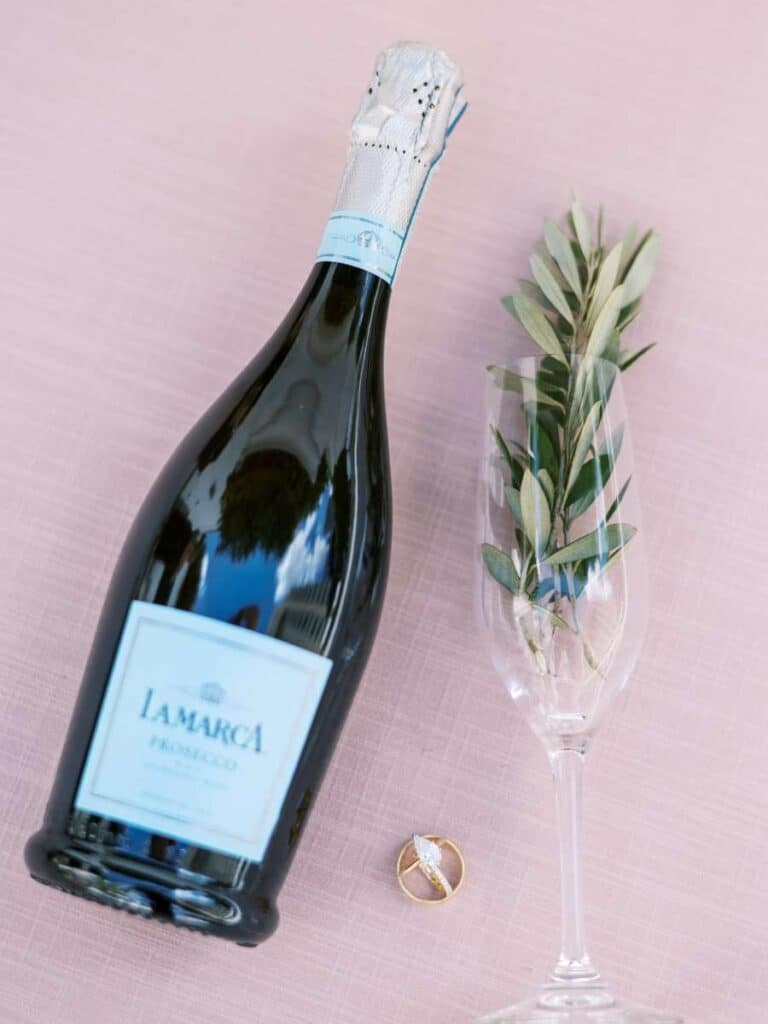 bottle of la marca prosecco with a greenery garnished glass and wedding rings